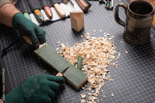 Carving tools, sharping knife on a leather strop. Cutting mat with basswood, shavings, gloves, knives, and coffee mug in background.
