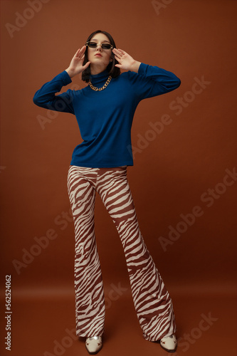 Fashionable confident woman wearing trendy brown rectangular sunglasses, stylish blue turtleneck, flared trousers with zebra print, boots. Full-length studio portrait photo