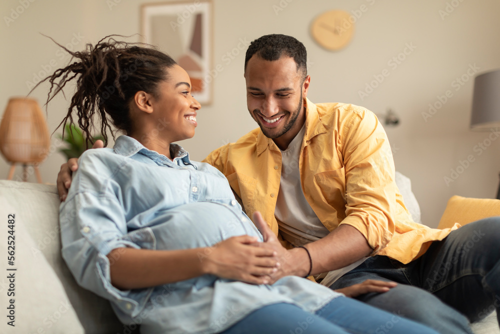 Obraz premium Happy african american couple expecting baby, pregnant black spouses embracing on couch at home