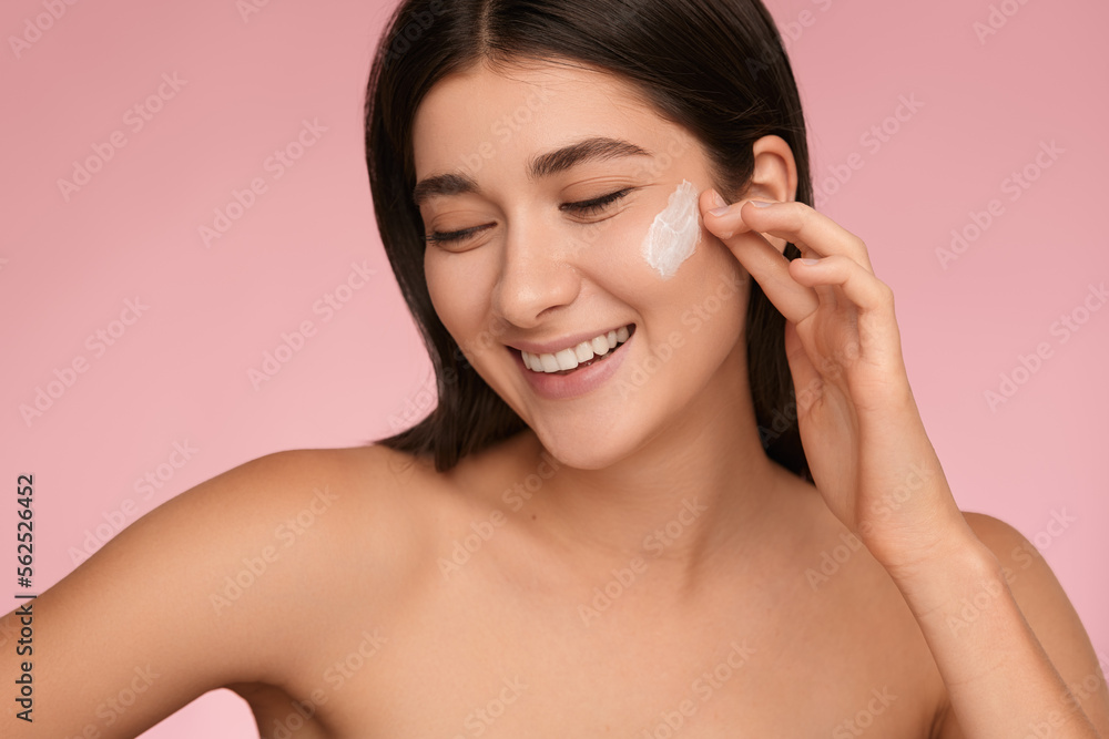 Smiling young lady applying moisturizing cream on face in pink studio