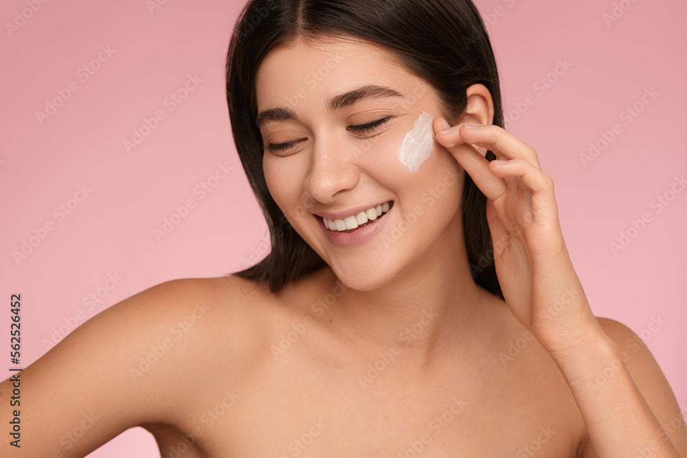 Smiling young lady applying moisturizing cream on face in pink studio Smiling young lady applying moisturizing cream on face in pink studio
