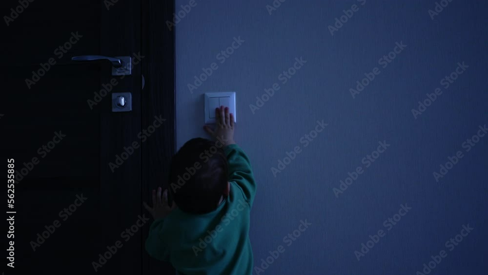 Small baby boy comes up to the light switch in the dark room. Beautiful toddler turns on the light and walks away happily.