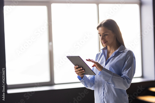 Confident business expert. Attractive young woman in smart casual wear holding digital tablet and looking at camera while leaning on desk in creative office