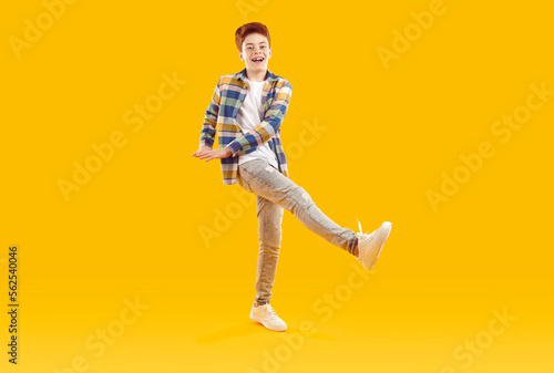 Happy funny redhead teenage boy raised up his leg. Full length shot of attractive joyful boy in casual plaid shirt and jeans standing on one leg over isolated yellow studio background