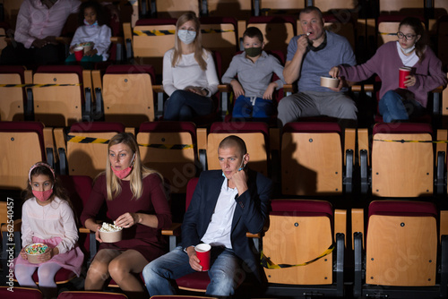 Group of people of different ages and nationalities wearing protective face masks to prevent viral infections absorbedly watching movie in cinema