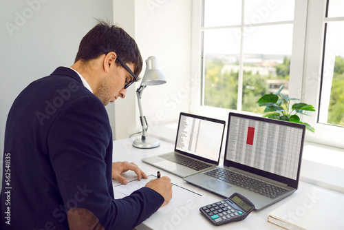 Financial accountant working in modern office. Busy man in suit and glasses sitting at desk by window, using calculator and two laptop computers, working with digital sheets and taking notes on paper © Studio Romantic