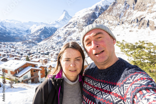 Cheerful adult man and woman enjoying travel in Swiss Alps in wintertime, making selfie together against snow covered mountain range and small town in foothills on sunny day