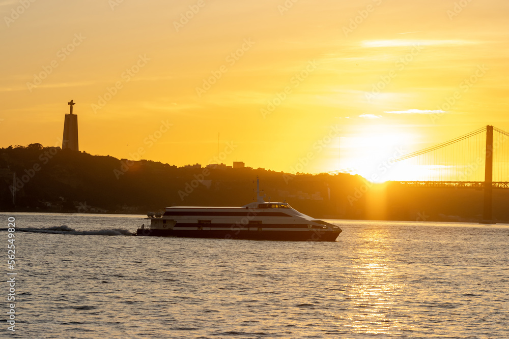 A yacht sails along the Tagus River at an orange sunset - a statue of Jesus Christ in the distance