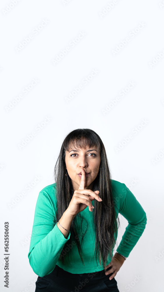 Brown hair woman with Hispanic features making silence gesture with her finger on lips over isolated white background,. Vertical photography with copy space above Brown hair woman with Hispanic features making silence gesture with her finger on lips over isolated white background,. Vertical photography with copy space above