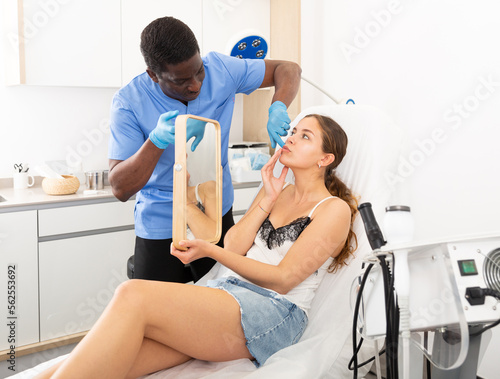 Professional cosmetologist examining female client face before procedure in esthetic clinic. Woman looking at mirror