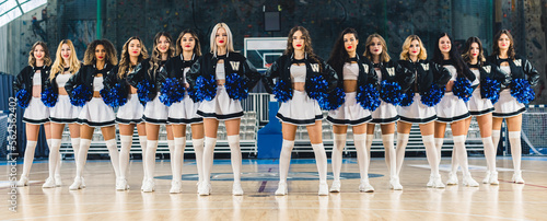 Front view of cheerleaders in uniforms standing in row and holding blue shiny pom-poms. Basketball court in the background. High quality photo