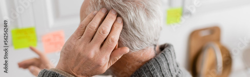 aged man with alzheimer disease touching head and pointing at blurred sticky notes in kitchen, banner.