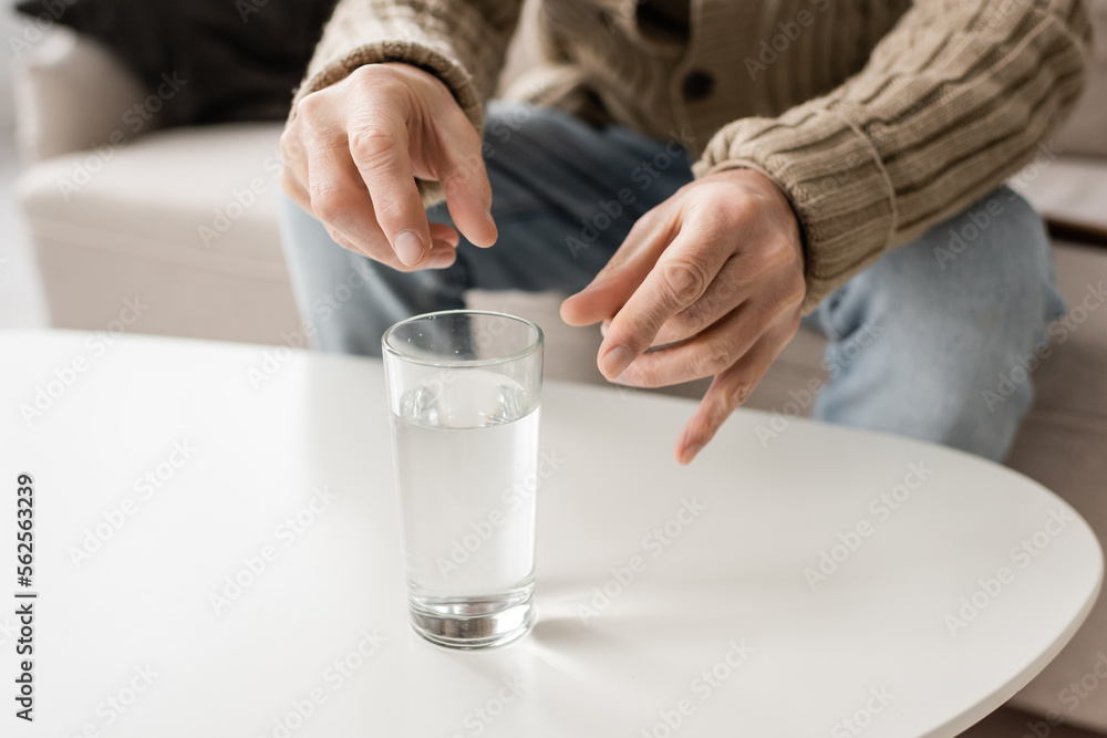 cropped view of man with parkinson disease and trembling hands sitting near glass of water at home.