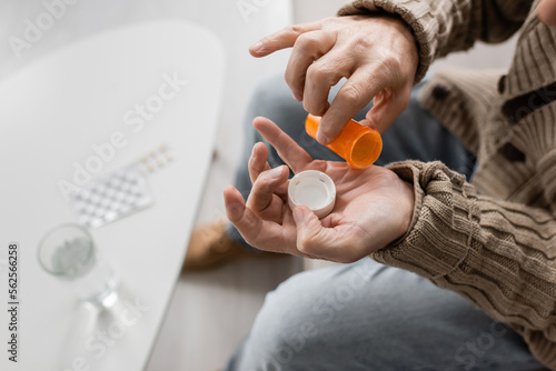 partial view of man with parkinson disease and tremor in hands holding pills container near blurred glass of water.