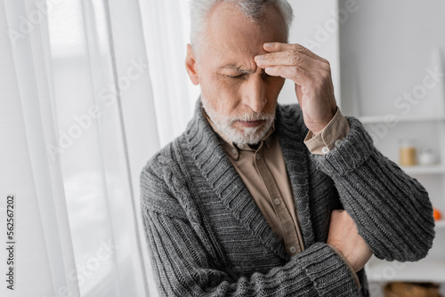 depressed senior man with alzheimer syndrome standing with closed eyes and touching forehead at home.