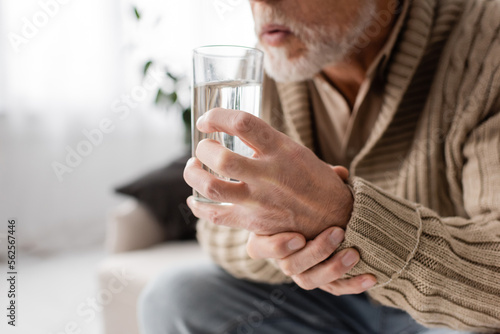 cropped view of aged man with parkinsonism holding glass of water in trembling hands while sitting at home. photo