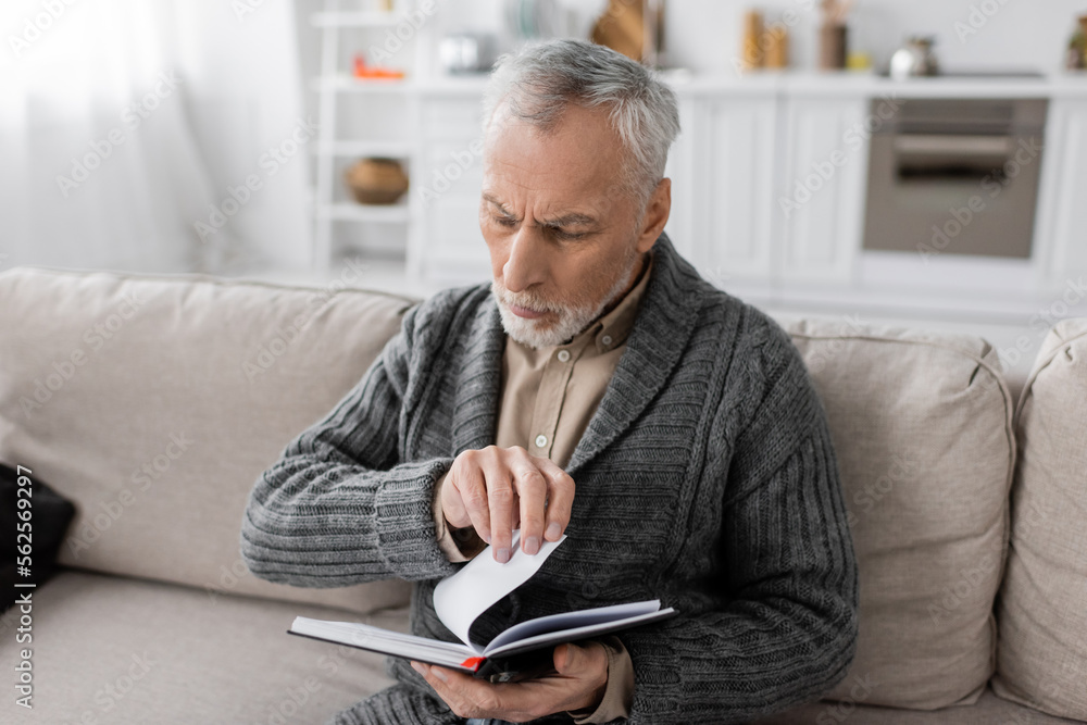 grey haired man suffering from memory loss caused by alzheimer disease looking in notebook on couch at home. grey haired man suffering from memory loss caused by alzheimer disease looking in notebook on couch at home.