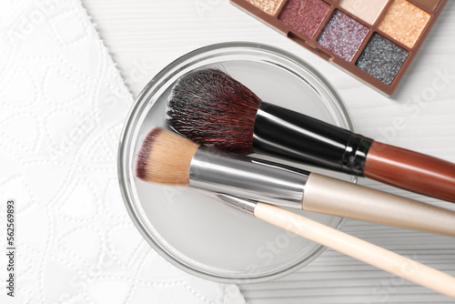 Cleaning makeup brushes in bowl with special liquid on white wooden table, flat lay