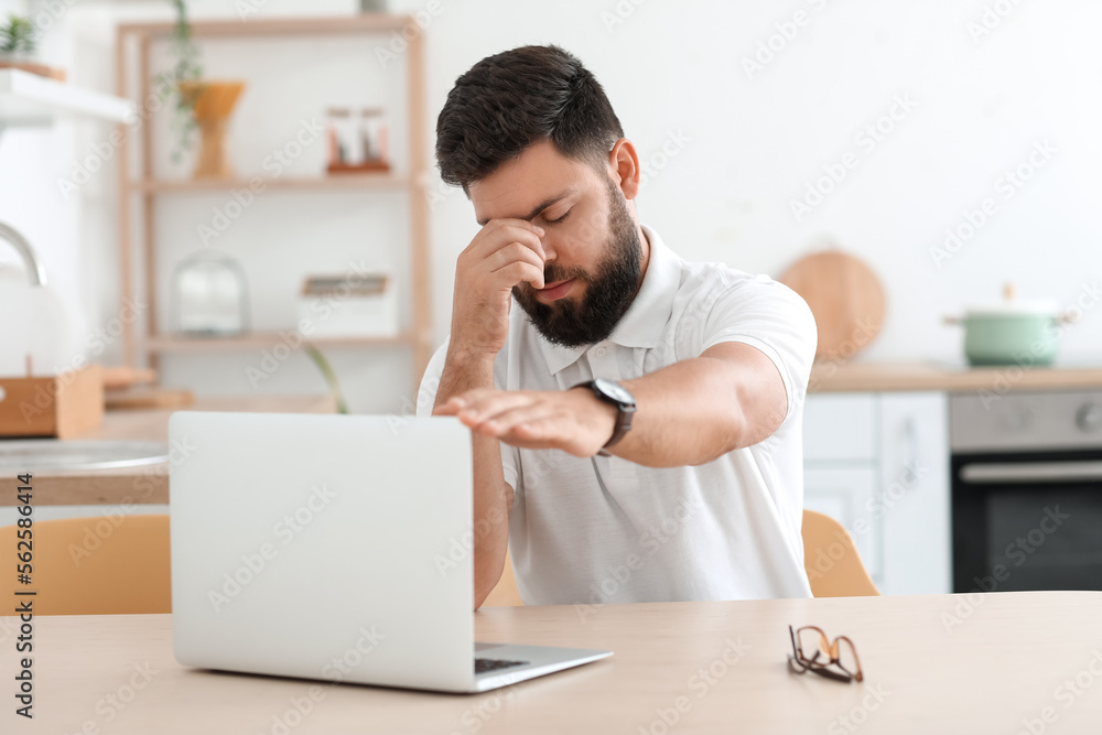 Thoughtful young bearded man with laptop at table in kitchen Thoughtful young bearded man with laptop at table in kitchen