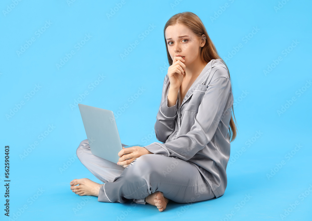 Young woman in pajamas with laptop biting nails on blue background Young woman in pajamas with laptop biting nails on blue background