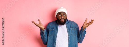 Confused african-american man in beanie looking at upper left corner doubtful, shrugging uncertain, standing over pink background photo