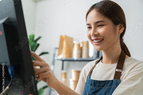 Happy young Asian woman cashier wears an apron and using pos terminal to input orders on coffee shop counter.