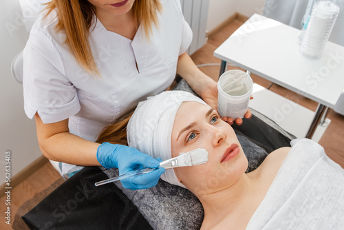 Hands of cosmetology specialist applying facial mask using brush making skin hydrated and face glowing and skin. Relaxed young female is resting on couch while cosmetologist massaging her face.