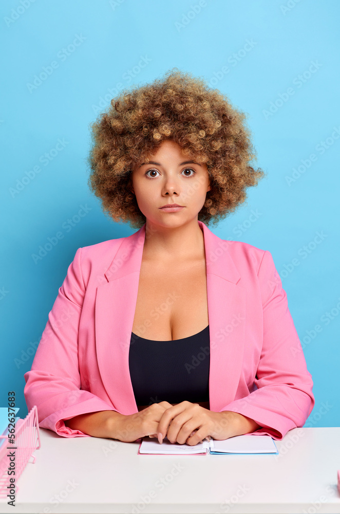 Pretty curly haired woman employee recruiter, seats at the white office desk with a normal come emotion, isolated over blue wall Pretty curly haired woman employee recruiter, seats at the white office desk with a normal come emotion, isolated over blue wall