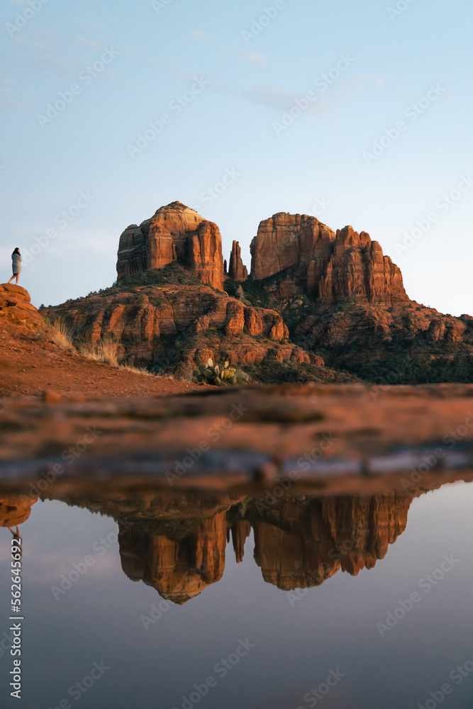 Young beautiful woman standing on rock point looking out at Cathedral Rock in Sedona Arizona USA Southwest at sunset with reflection from small pool.