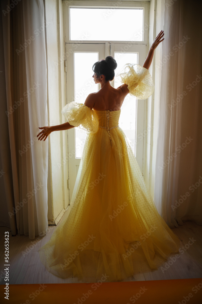 Fototapeta premium A woman's silhouette in a golden luxurious dress against the background of a window holds a curtain with her hands. Elegant lady in a yellow long silk dress with bare back, back view.