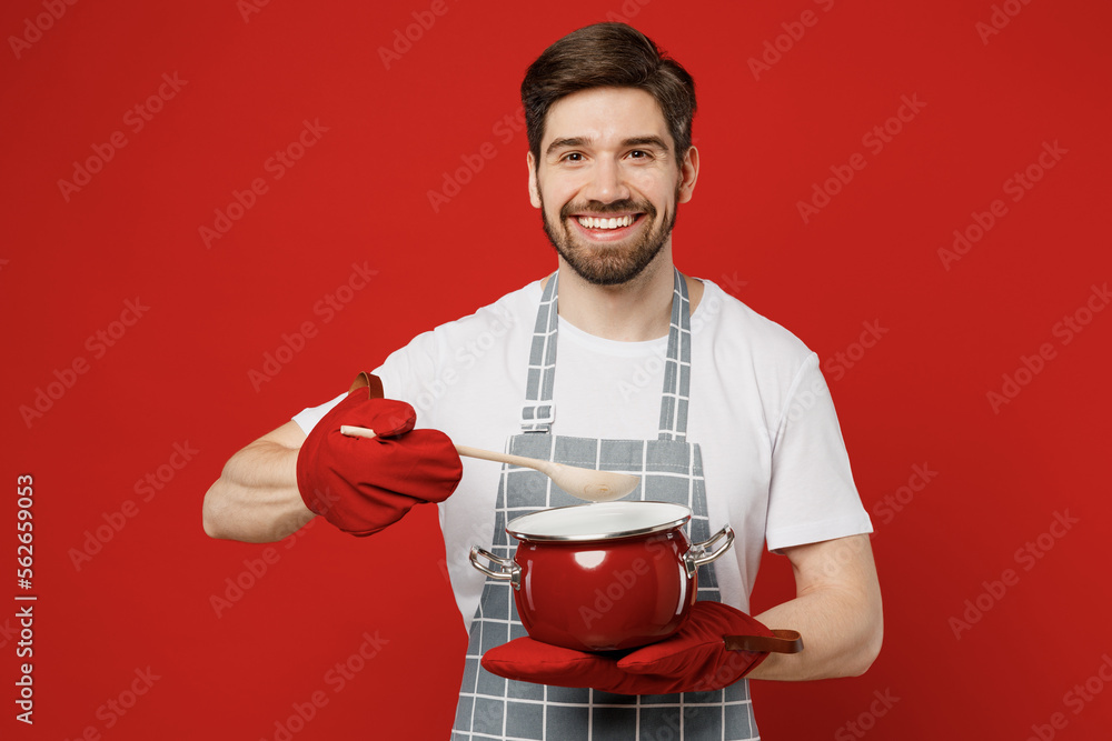 Young smiling happy cheerful male housewife housekeeper chef cook baker man in grey apron oven mittens hold saucepan spoon looking camera isolated on plain red background studio Cooking food concept Young smiling happy cheerful male housewife housekeeper chef cook baker man in grey apron oven mittens hold saucepan spoon looking camera isolated on plain red background studio Cooking food concept