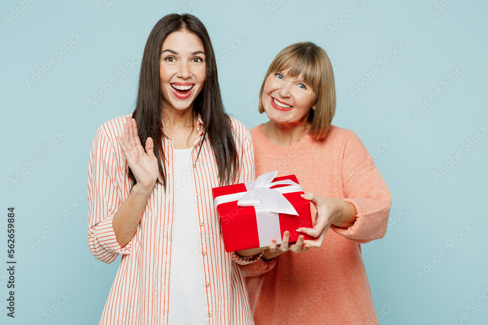 Surprised elder parent mom with young adult daughter two women together wear casual clothes hold in hands present box with gift ribbon bow isolated on plain blue cyan background. Family day concept. Surprised elder parent mom with young adult daughter two women together wear casual clothes hold in hands present box with gift ribbon bow isolated on plain blue cyan background. Family day concept.