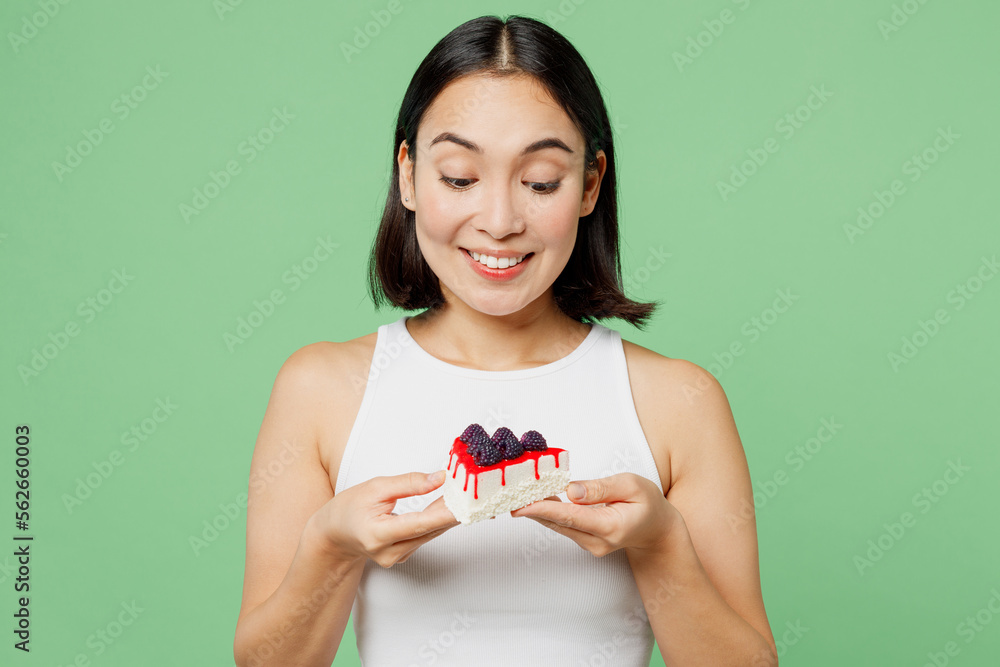 Young smiling happy fun woman wearing white clothes holding in hand pice of cake dessert isolated on plain pastel light green background. Proper nutrition healthy fast food unhealthy choice concept. Young smiling happy fun woman wearing white clothes holding in hand pice of cake dessert isolated on plain pastel light green background. Proper nutrition healthy fast food unhealthy choice concept.