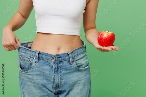 Cropped young woman wears white clothes show loose pants on waist after weightloss hold red apple isolated on plain light green background. Proper nutrition healthy fast food unhealthy choice concept.