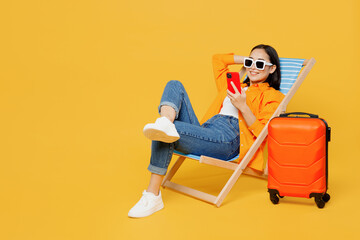 Young fun woman wear summer clothes sit in deckchair use mobile cell phone isolated on plain yellow background. Tourist travel abroad in free spare time rest getaway. Air flight trip journey concept.