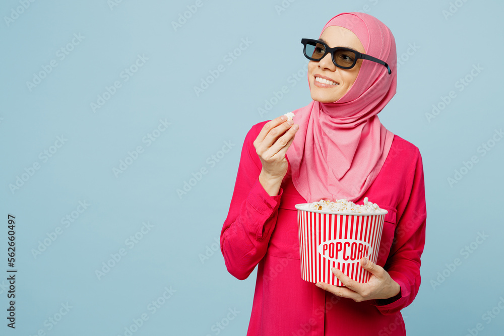 Obraz premium Young smiling happy arabian muslim woman woman in 3d glasses abaya hijab watching movie film hold bucket of popcorn in cinema look aside on area isolated on plain blue cyan background studio portrait.