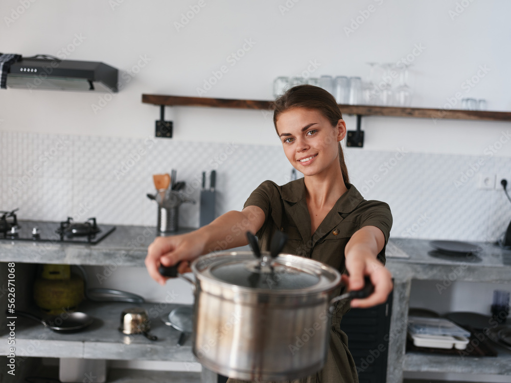 A woman in stylish kitchen prepares food and holds a cooking pot in her hands for cooking dinner, camera and smiles with teeth, looking into the camera. Home atmosphere, lifestyle, comfort A woman in stylish kitchen prepares food and holds a cooking pot in her hands for cooking dinner, camera and smiles with teeth, looking into the camera. Home atmosphere, lifestyle, comfort