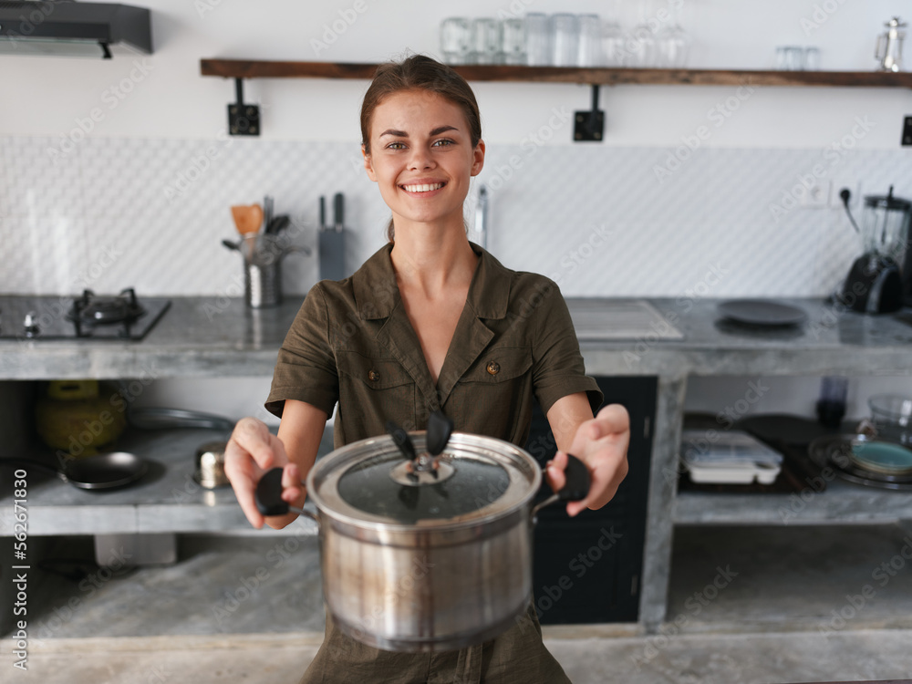 A woman in stylish kitchen prepares food and holds a cooking pot in her hands for cooking dinner, camera and smiles with teeth, looking into the camera. Home atmosphere, lifestyle, comfort A woman in stylish kitchen prepares food and holds a cooking pot in her hands for cooking dinner, camera and smiles with teeth, looking into the camera. Home atmosphere, lifestyle, comfort