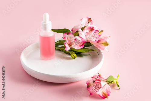 A beautiful cosmetic bottle with a white pipette stands on a ceramic plate with astromeria flowers. pink background. natural cosmetics.