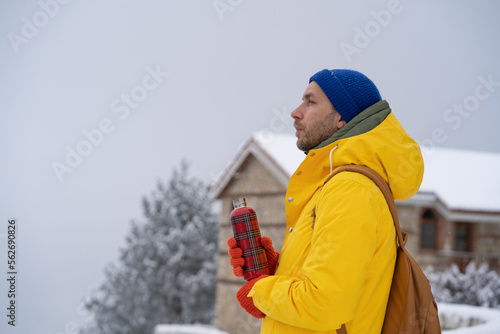 Thoughtful man in warm clothes with backpack left house with thermos in cold snowy weather with frosty air looks lost in thoughts. Confident European male stands outdoor looks away, dreams of future.