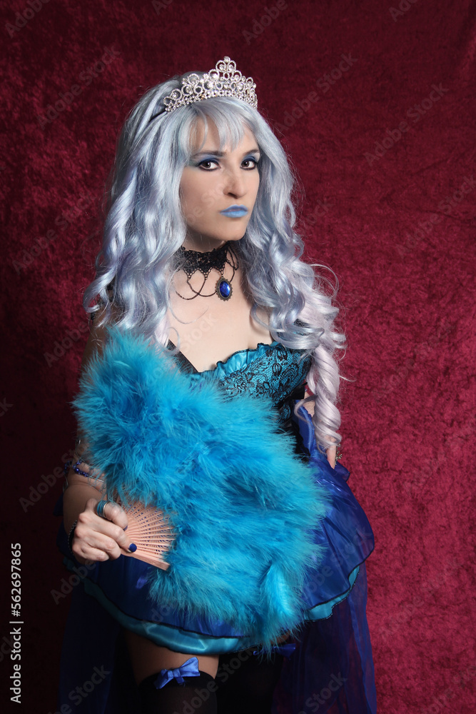 Fototapeta premium Woman With Blue Hair and Blue Corset Dress Holding Blue Fan on Red Velvet Background