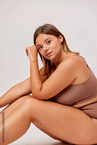Vertical shot of body positive confident young woman sits on a floor in studio in beige lingerie, isolated over white background 