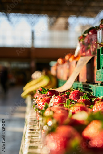 Strawberries on a stand inside of a farmers market in close-up, bananas blurred in the background. 