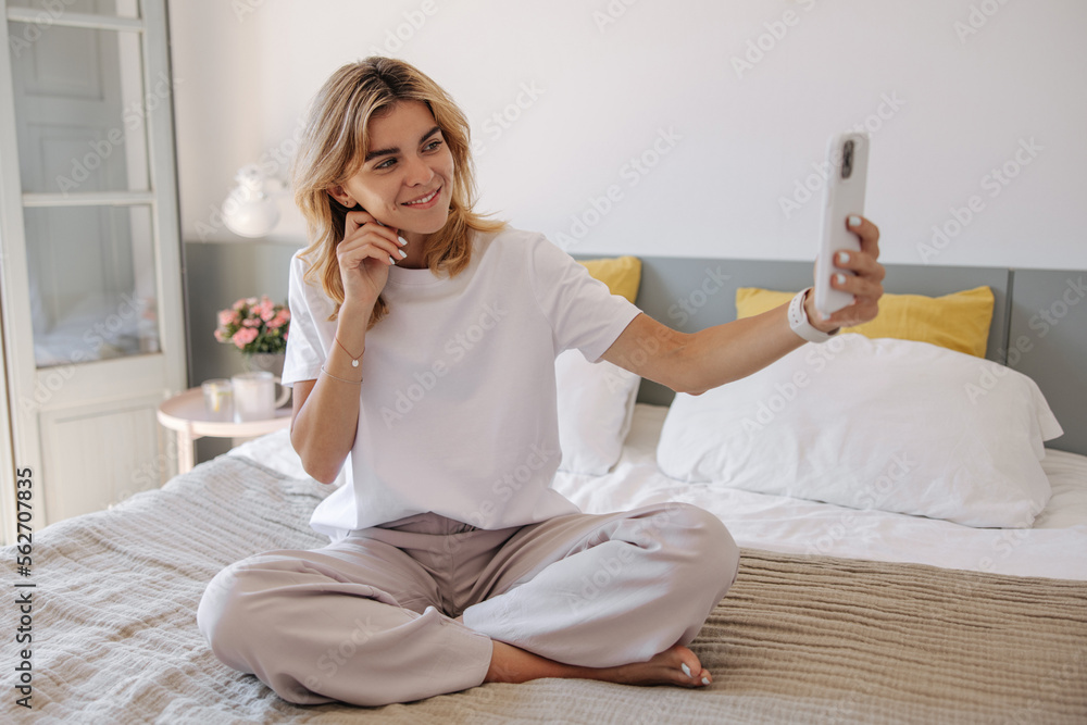 Cute young caucasian girl takes selfie with smartphone sitting on bed in morning. Blonde hair woman wears t-shirt and casual pants. Concept gadgets Cute young caucasian girl takes selfie with smartphone sitting on bed in morning. Blonde hair woman wears t-shirt and casual pants. Concept gadgets