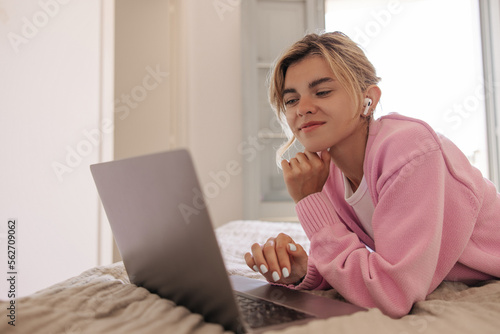 Pretty young caucasian female dressed in pink sweater lying and working on bed at home. Calm woman blonde hair with good mood uses laptop. Concept technologies  lifestyle.