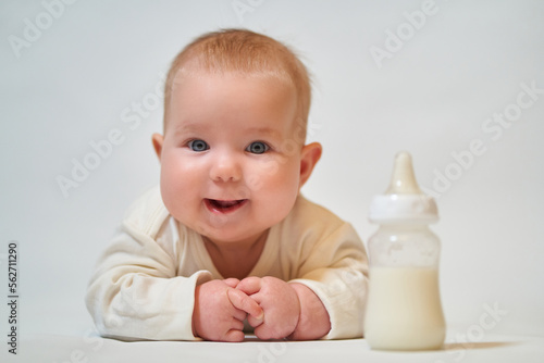 portrait of a smiling baby in light clothes next to a bottle of milk on a light background