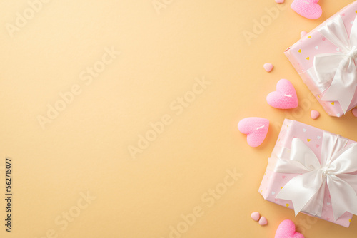 Valentine's Day concept. Top view photo of gift boxes with white ribbon bows heart shaped candles and sprinkles on isolated pastel beige background with copyspace