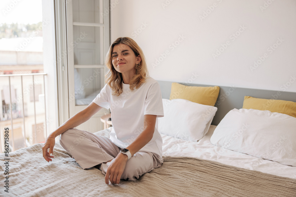 Cute young caucasian girl with smart watch sitting on bed near panoramic window at home. Blonde hair woman dressed in white t-shirt and casual pants. Concept enjoying morning, lifestyle. Cute young caucasian girl with smart watch sitting on bed near panoramic window at home. Blonde hair woman dressed in white t-shirt and casual pants. Concept enjoying morning, lifestyle.