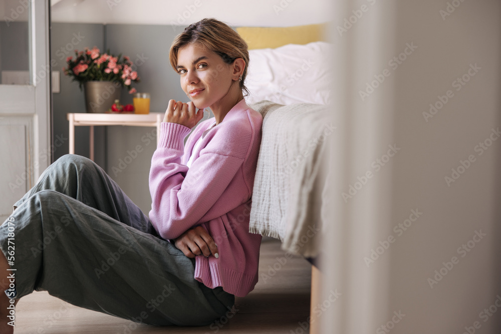 Pretty young caucasian girl dressed in casual clothes sitting on floor near bed in morning. Blonde hair woman relaxing alone at room. Concept privacy home. Pretty young caucasian girl dressed in casual clothes sitting on floor near bed in morning. Blonde hair woman relaxing alone at room. Concept privacy home.