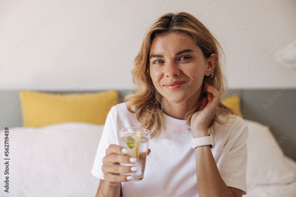 Pretty young caucasian girl blonde holds glass of water with slice of lemon sitting on bed. Smiling woman with smart watch looking at camera. Concept positive emotions, lifestyle. Pretty young caucasian girl blonde holds glass of water with slice of lemon sitting on bed. Smiling woman with smart watch looking at camera. Concept positive emotions, lifestyle.